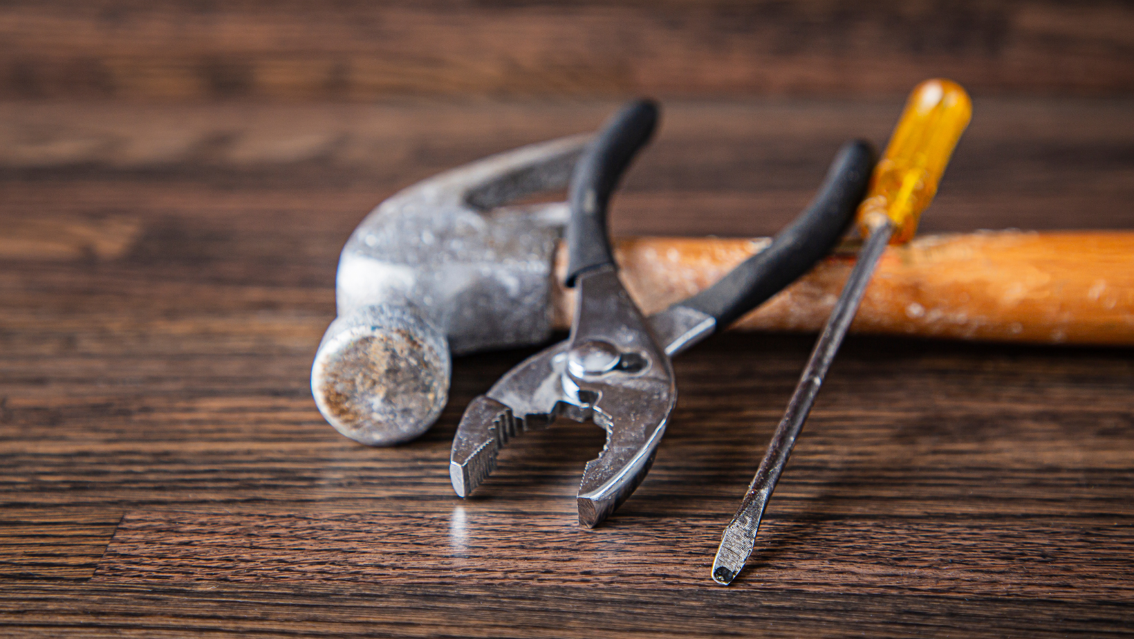 hammer, screwdriver and pliers laying against a wood backdrop